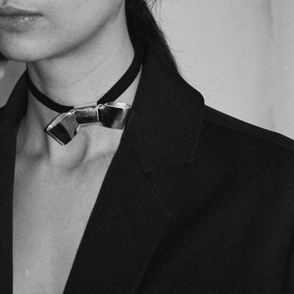 BOW FAUX SUEDE RIBBON CRAVAT NECKLACE featuring a clean chic design and modern nature silver and black color ,perfect for dailylife/party/office/casual occasion. Displayed in a lifestyle setting with a model wearing a black  blazer.