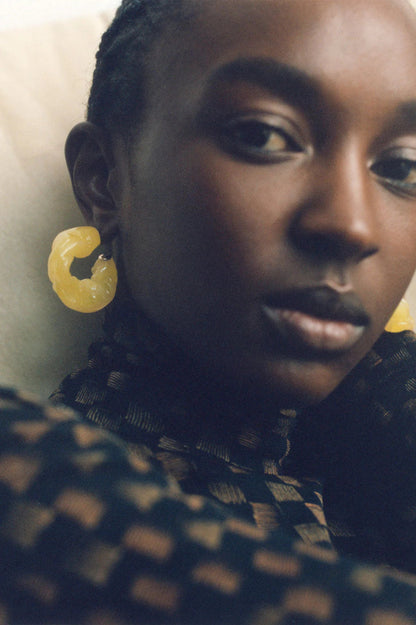 CULT STATEMENTPIECES BLUE BIO RESIN GOLD VERMEIL EARRINGS featuring a clean chic hoop design and modern jelly green color ,perfect for dailylife/party/office/casual occasion. Displayed in a cool  lifestyle setting with a black skin model wearing a black pants.