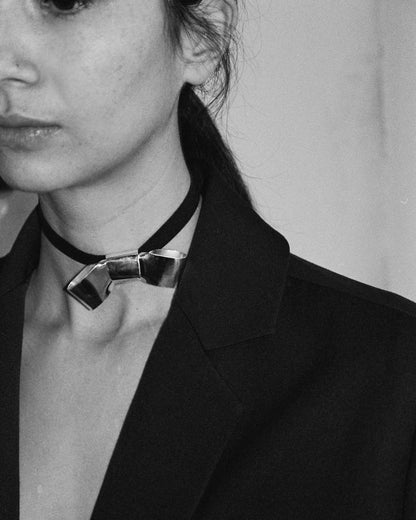 BOW FAUX SUEDE RIBBON CRAVAT NECKLACE featuring a clean chic design and modern nature silver and black color ,perfect for dailylife/party/office/casual occasion. Displayed in a lifestyle setting with a model wearing a black  blazer.